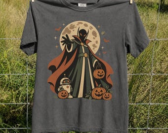 May include: A charcoal gray t-shirt featuring a graphic print of a caped figure in a dark robe with orange accents, standing before a large full moon. The figure is surrounded by three jack-o'-lanterns and a small robot, with bats and stars.