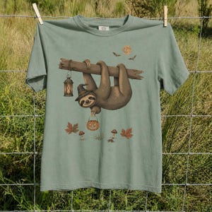 May include: A sage green t-shirt featuring a graphic of a brown sloth wearing a witch hat, hanging from a branch. The sloth holds a jack-o'-lantern bucket, with a hanging lantern, a full moon, bats, autumn leaves, and mushrooms printed below.