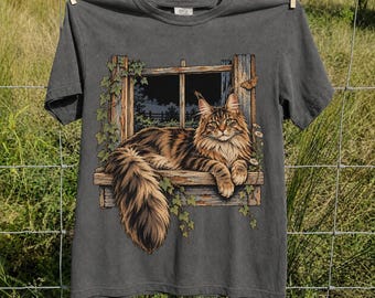 May include: A charcoal gray short-sleeve t-shirt featuring a graphic print of a brown tabby cat resting on a wooden window sill. The window frame is adorned with green ivy vines and a small butterfly, set against a dark night sky background.