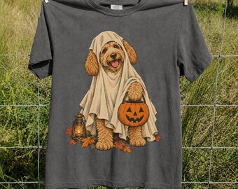 May include: A charcoal gray short-sleeve t-shirt featuring a graphic of a golden-colored dog wearing a white ghost costume. The dog holds an orange jack-o'-lantern bucket, stands among fallen autumn leaves, and is next to a lantern.