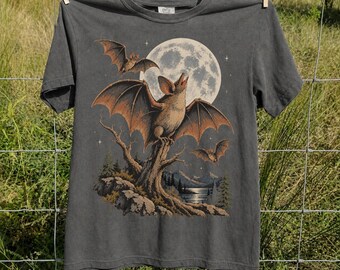May include: A charcoal gray short-sleeve t-shirt featuring a graphic print of three bats flying and perched on a tree branch against a large full moon. The illustration includes a landscape with mountains, a lake, and pine trees in muted earth tones.