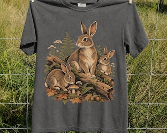 May include: A charcoal gray short-sleeve t-shirt featuring a graphic print of three brown rabbits resting on a fallen log. The scene includes green ferns, small mushrooms, and scattered autumn leaves surrounding the rabbits in a forest setting.