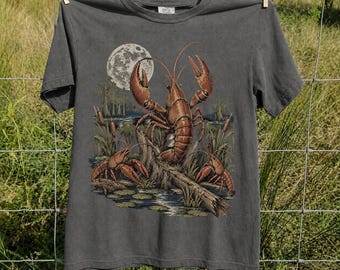 May include: A charcoal gray short-sleeve t-shirt featuring a graphic print of three brown lobsters in a swampy landscape with cattails, lily pads, and a large full moon in the background. The design is centered on the front of the garment.