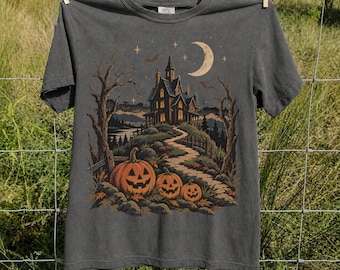 May include: A charcoal gray short-sleeve t-shirt featuring a graphic print of a haunted house on a hill, a crescent moon, bats, and three jack-o'-lanterns in the foreground. The design includes bare trees and a winding path leading to the house.