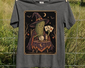 May include: A charcoal gray t-shirt featuring a graphic of a pickle wearing a witch hat, sitting at a table with a crystal ball, a candle, and three tarot cards. The illustration includes a crescent moon, stars, and bats against a dark background.