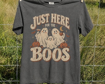 May include: A charcoal gray short-sleeve t-shirt featuring a graphic print of three white ghosts, two orange pumpkins, and the text "JUST HERE FOR THE BOOS" in a vintage-style font. The design includes small stars and a crescent moon accent.