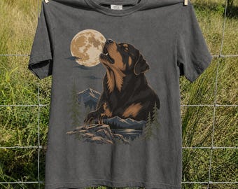 May include: A charcoal gray short-sleeve t-shirt featuring a graphic print of a Rottweiler dog howling at a full moon. The illustration includes a smaller dog standing on a rocky cliff, pine trees, and distant mountains under a night sky.
