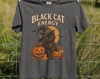 May include: A charcoal gray short-sleeve t-shirt featuring a graphic print of a black cat wearing a witch hat, positioned between two orange jack-o'-lanterns and autumn leaves. The text "BLACK CAT ENERGY" is printed in a curved font above.