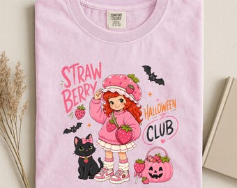 May include: A light pink t-shirt featuring a graphic of a red-haired character in a strawberry hat and sweater, a black cat with a pink bow, two bats, and a pink pumpkin bucket. The text reads STRAWBERRY HALLOWEEN CLUB in pink and orange.