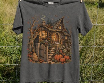 May include: A charcoal gray short-sleeve t-shirt featuring a graphic print of a cottage with a stone path, pumpkins, a broom, and a black cat. The scene includes autumn foliage, a chimney with smoke, and a night sky with stars and bats.