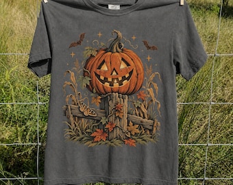 May include: A charcoal gray short-sleeve t-shirt featuring a graphic print of a carved jack-o'-lantern pumpkin sitting on a wooden fence post, surrounded by autumn leaves, corn stalks, a moth, and two flying bats under small star icons.