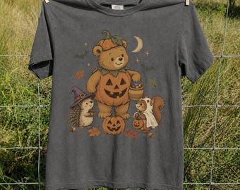 May include: A charcoal gray t-shirt featuring a graphic illustration of a bear wearing a pumpkin costume, a hedgehog in a purple witch hat, and a squirrel dressed as a ghost. The design includes a crescent moon, stars, bats, and autumn leaves.