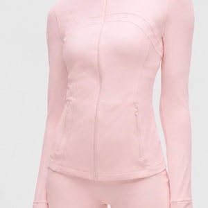 Op de afbeelding: Een lichtroze jas met lange mouwen en ritssluiting, voorzien van een hoge kraag en twee voorzakken met rits, gecombineerd met bijpassende lichtroze shorts. De jas heeft getailleerde naden en lange mouwen met duimgaten in de boorden.