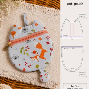 May include: A light blue fabric pouch shaped like a cat head with a fox and berry print, featuring a peach-colored zipper, a small fabric loop, and a tail-shaped tab. The image includes a PDF sewing pattern diagram labeled cat pouch.