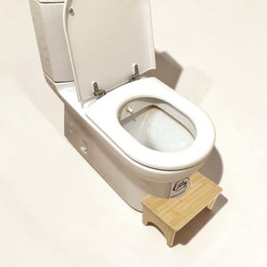 May include: A white ceramic toilet with an open lid and seat, featuring a light wood-toned rectangular footstool positioned directly in front of the base. The footstool has a small white sticker with the text Ealy printed on the front side.