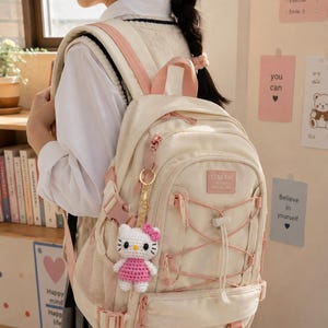 May include: A cream-colored backpack with light pink accents, featuring a front pocket with a drawstring cord, side mesh pockets, and adjustable straps. A small crocheted cat figure in a pink dress hangs from a gold-toned metal keychain.