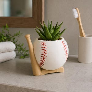 Peut inclure: Un pot de fleurs en céramique blanche en forme de balle de baseball avec des coutures rouges, soutenu par un support en bois clair en forme de batte de baseball. Une plante succulente verte est plantée à l'intérieur de la balle.