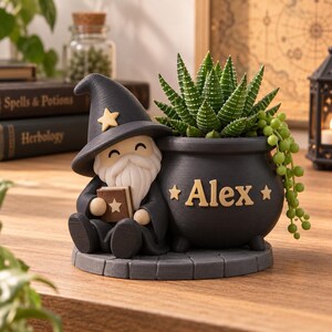 Könnte beinhalten: Ein schwarzer Übertopf im Zauberer-Design mit einem sitzenden Gnom mit weißem Bart und spitzem Hut, der ein kleines Buch hält. Der angebrachte kesselförmige Topf zeigt den Namen Alex in goldener Schrift, flankiert von zwei goldenen Sternen.