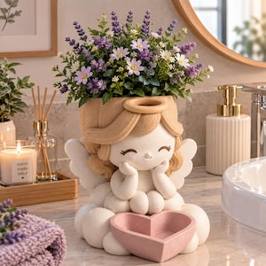 May include: A cream-colored angel-shaped planter featuring a smiling face, blonde hair, and wings, resting on a cloud base. The angel holds a pink heart-shaped bowl in front. The planter is filled with a bouquet of purple lavender and white daisies.