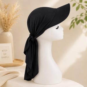 May include: A solid black head covering featuring a built-in visor and a knotted back detail. The fabric wraps around the head, securing with a knot at the nape of the neck, with the remaining material draping downwards to provide coverage.