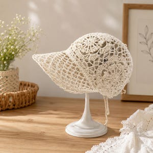May include: A cream-colored crochet hat featuring an intricate floral pattern on the crown and a wide, structured brim. The design is crafted from fine yarn with an openwork mesh texture, including delicate hanging strings at the back.