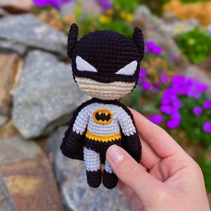Peut inclure: Une petite poupée crochetée à la main représentant un super-héros, dotée d'une cagoule noire à oreilles pointues, d'yeux blancs, d'un visage beige, d'un torse gris avec un emblème jaune et noir, d'une ceinture jaune et d'une cape noire.