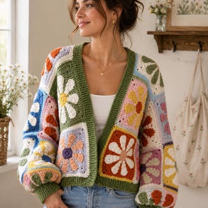 May include: A long-sleeved crochet cardigan featuring a patchwork design of multi-colored floral squares. The garment has a deep olive green ribbed trim along the front opening, cuffs, and hem, with various flower motifs in white, pink, blue, and orange.