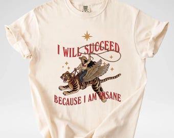 May include: A cream-colored short-sleeve t-shirt featuring a graphic of a person in a cowboy hat riding a winged tiger while holding a lasso. The text I WILL SUCCEED BECAUSE I AM INSANE is printed in red above and below the central illustration.