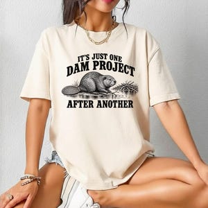 May include: A cream-colored short-sleeve t-shirt featuring a black graphic of a beaver near a dam. The text printed in black above and below the beaver reads, IT'S JUST ONE DAM PROJECT AFTER ANOTHER. The shirt has a crew neckline and a relaxed fit.
