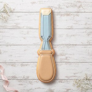 May include: A chisel-shaped biscuit decorated with royal icing. The blade section is light blue with a cream-coloured tip, while the handle is a solid tan colour. The icing features defined outlines and smooth surfaces, mimicking the appearance of a tool.