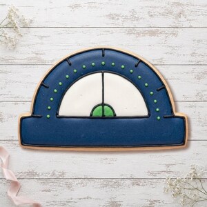 Peut inclure: Un biscuit en forme de demi-cercle représentant un rapporteur, décoré avec un glaçage bleu foncé, un centre blanc et de petits points verts le long de l'arc. Le motif présente des contours en glaçage noir et un petit demi-cercle vert à la base.
