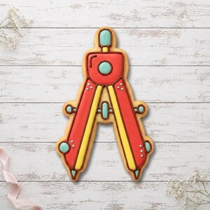 Peut inclure: Un biscuit en forme de compas décoré avec du glaçage royal aux couleurs rouge vif, jaune éclatant et bleu clair. Le motif présente un corps central rouge avec des accents jaunes, des détails circulaires bleu clair et des contours bruns.