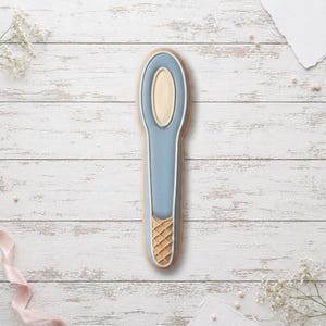 May include: A biscuit shaped like a spoon with a light blue handle, a cream-coloured oval bowl, and a tan waffle-textured pattern at the base of the handle. The biscuit features clean white piping outlines that define the spoon's structural shape.