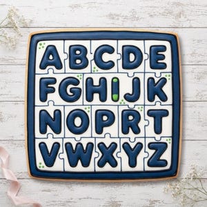 May include: A square biscuit decorated with white icing and dark blue letters of the alphabet arranged in a grid of puzzle pieces. The letters A through Z are present, with a small green accent on the letter I. The biscuit has a dark blue border.