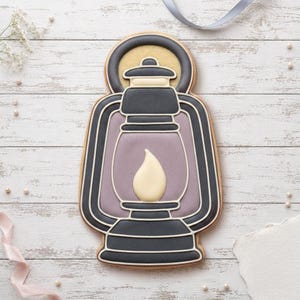 May include: A lantern-shaped biscuit decorated with dark grey icing for the frame and handle, a light purple centre panel, and a cream-coloured flame detail. The design features clean white piped outlines defining the structure of the oil lamp.