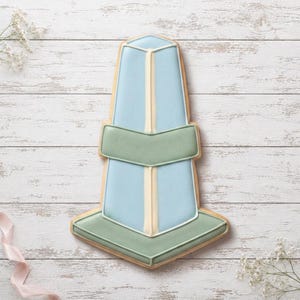May include: A sugar cookie shaped like a nautical buoy, decorated with smooth light blue and sage green icing. The design features a vertical cream-colored stripe running down the center, with horizontal sage green bands across the middle and base.