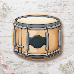 May include: A drum-shaped biscuit decorated with tan icing for the shell and grey icing for the rims, tension rods, and a central badge. The design features a smooth, rounded top surface and detailed piping to replicate the hardware of a snare drum.