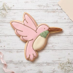 May include: A hummingbird-shaped sugar biscuit decorated with smooth pink icing on the wings and body, a cream-coloured belly, a textured sage green throat patch, and a brown beak, featuring piped outlines defining the bird's form.