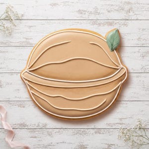 May include: A round, tan-coloured biscuit decorated with smooth icing. The surface features raised, off-white piped lines creating a textured, layered pattern. A single, small, sage-green icing leaf is attached to the upper right edge of the biscuit.