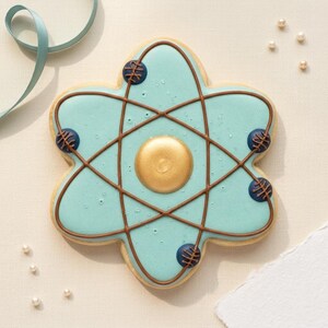 May include: A sugar cookie shaped like an atom with light blue icing, a central gold metallic circle, and dark blue circular accents. Thin brown icing lines form orbital paths around the center, decorated with small white sugar crystals.