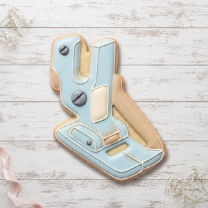 May include: A sugar biscuit shaped like a futuristic blaster or tool, decorated with light blue, cream, and grey icing. The design features raised piping details, including circular grey accents that resemble screw heads, set against a light tan biscuit base.