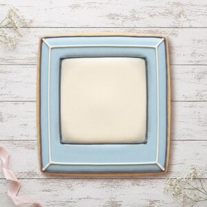 May include: A square-shaped cookie decorated with royal icing to resemble a picture frame. The center is a smooth, off-white square bordered by a dark grey line, surrounded by a wide, light blue border with thin white piping accents.