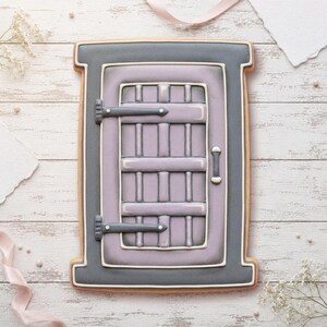 May include: A rectangular cookie decorated with royal icing to resemble a window. The frame is dark gray with white piping, while the central shutter is light purple with a grid pattern, dark gray hinges, and a small vertical handle on the side.