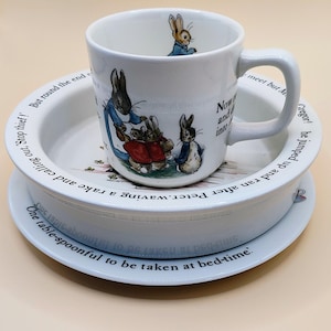 May include: A white ceramic mug and bowl set featuring Peter Rabbit illustrations and black printed text. The mug shows Peter Rabbit in a blue jacket, while the bowl rim displays the text: One table-spoonful to be taken at bed-time.