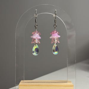 Può includere: Un paio di orecchini pendenti con ganci in metallo color argento, ciascuno con una perla a forma di fiore traslucido rosa chiaro a strati, sormontata da un petalo rosa più piccolo e un ciondolo in vetro iridescente a goccia.