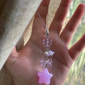 Può includere: Un ciondolo per telefono con una serie di perline color viola chiaro e traslucide attaccate a un anello in metallo argentato. Il ciondolo include una perlina a forma di quadrifoglio, una perla bianca, un fiore, un fiocco e una stella.