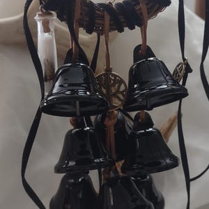May include: A decorative hanging ornament featuring a circular woven twig wreath wrapped in black velvet ribbon. Multiple glossy black metal bells are suspended from the wreath by brown leather cords, interspersed with a small glass vial and a metal tree charm.