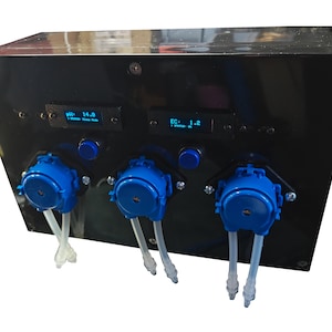 Könnte beinhalten: Eine schwarze, rechteckige Steuereinheit mit drei blauen Schlauchpumpen und transparenten Schläuchen. Die Frontplatte enthält zwei kleine Digitalanzeigen mit den Werten pH 14.0 und EC 1.2 sowie zwei blaue, runde Tasten über den Pumpen.