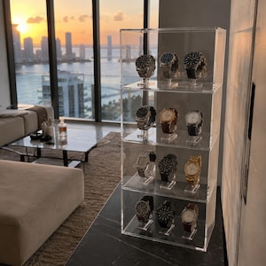 Peut inclure: Une vitrine en acrylique transparent à quatre étagères, contenant chacune trois montres-bracelets. La collection comprend diverses montres avec des bracelets en métal et en cuir dans des tons argent, or, noir et blanc, disposées sur des supports.