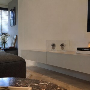 Pode incluir: Um expositor duplo de relógios em acrílico transparente contendo dois relógios de pulso prateados com mostradores pretos, posicionado sobre um móvel branco longo e suspenso na parede, ao lado de livros empilhados com as etiquetas Chanel e Dior.